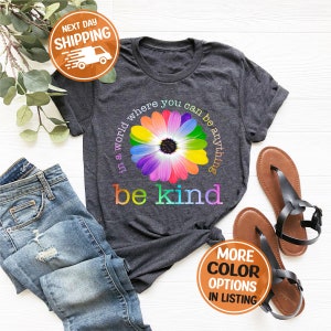 Puede incluir: Camiseta gris oscuro con un dise&ntilde;o de margarita colorida y la frase "in a world where you can be anything, be kind". La imagen tambi&eacute;n muestra vaqueros azules y sandalias marrones.