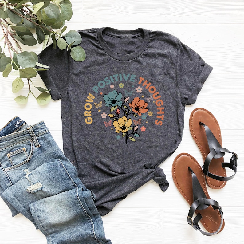 May include: A dark grey t-shirt with a floral design and the text "Grow Positive Thoughts" in a circle around the flowers.