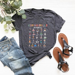 May include: A dark grey t-shirt with a colorful graphic that says "Coping Skills Alphabet" and features a list of coping skills for each letter of the alphabet.