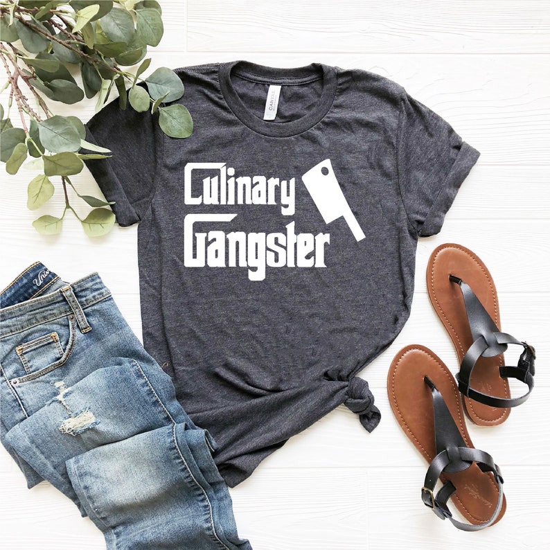 May include: A dark heather grey t-shirt with the text "Culinary Gangster" in white, featuring a white silhouette of a butcher knife.
