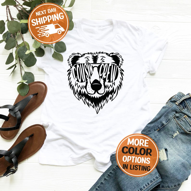 May include: White t-shirt featuring a black bear graphic wearing sunglasses that say "Cousin Crew".