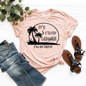 May include: A pink t-shirt with a black graphic that says "It's 5 o'clock everywhere I'm retired" with palm trees and a sunset.