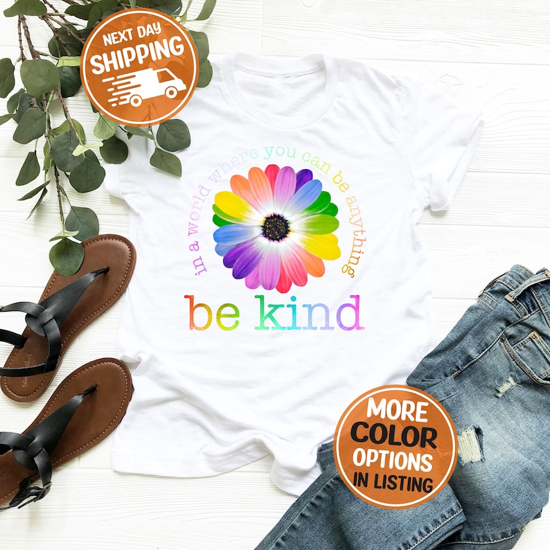 Puede incluir: Camiseta blanca con un dise&ntilde;o de margarita colorida y las palabras "in a world where you can be anything, be kind". Los p&eacute;talos de la margarita son de color arco&iacute;ris. La camiseta se combina con vaqueros azules y sandalias negras.