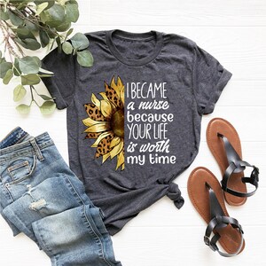 Op de afbeelding: Een grijs T-shirt met een zonnebloemdessin en de tekst "Ik ben verpleegster geworden omdat je leven mijn tijd waard is".