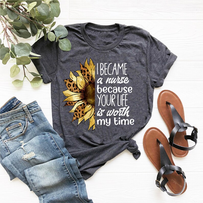 Op de afbeelding: Een grijs T-shirt met een zonnebloemdessin en de tekst "Ik ben verpleegster geworden omdat je leven mijn tijd waard is".