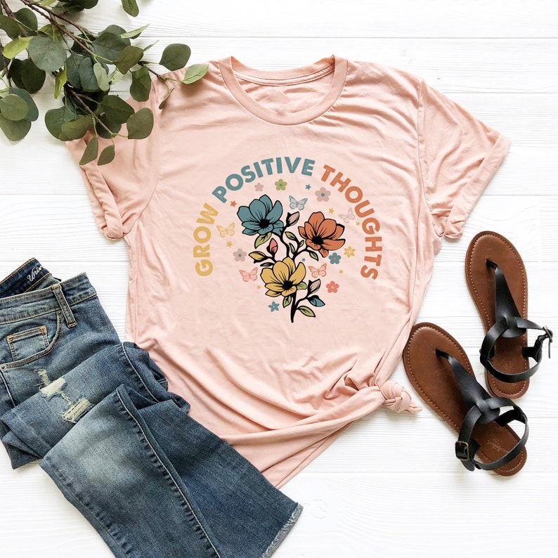 May include: A pink t-shirt with a graphic design of flowers and butterflies. The text "Grow Positive Thoughts" is written in a circle around the flowers.