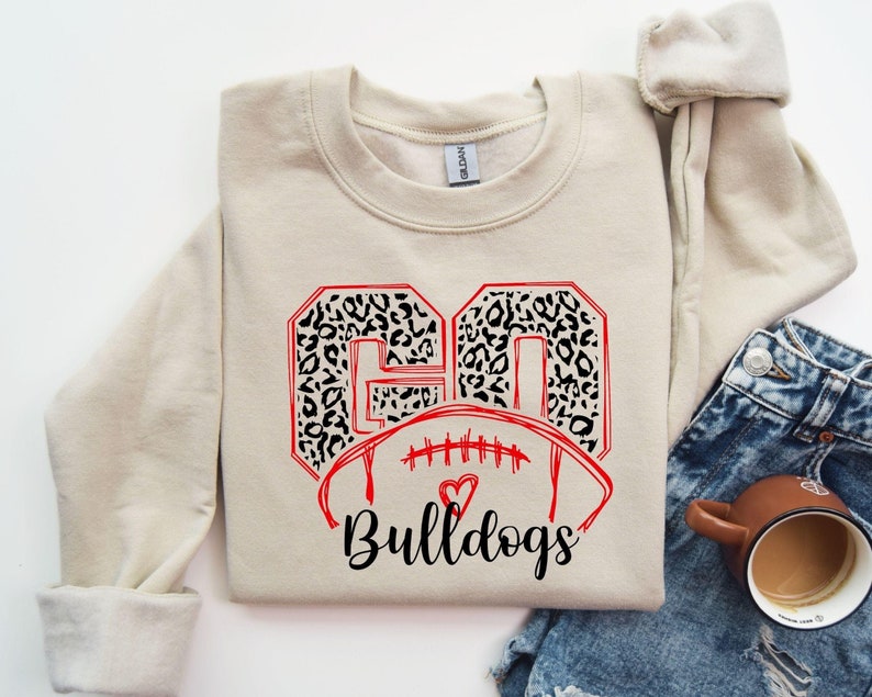May include: A beige sweatshirt with a red and black graphic design. The graphic features the numbers "60" in a leopard print, a football, and the text "Bulldogs".