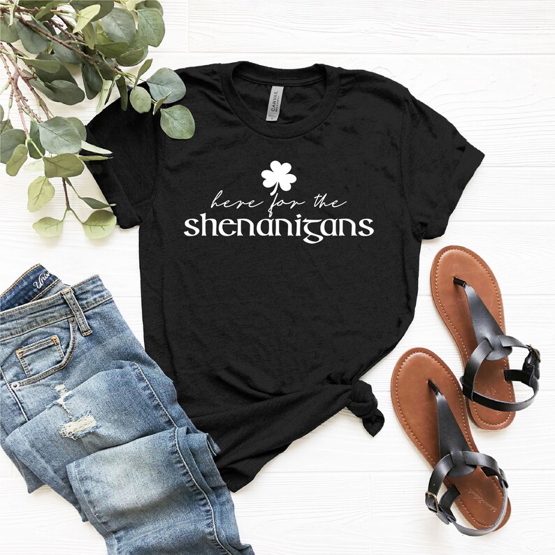May include: Black t-shirt with white text that reads "Here for the shenanigans" and a four-leaf clover graphic.