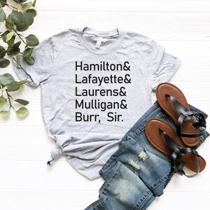 Op de afbeelding: Een grijs T-shirt met de tekst "Hamilton & Lafayette & Laurens & Mulligan & Burr, Sir." erop gedrukt. Het shirt ligt op een wit oppervlak met een paar bruine sandalen en een blauwe jeans.