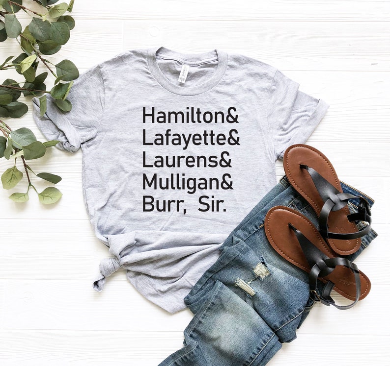 Op de afbeelding: Een grijs T-shirt met de tekst "Hamilton & Lafayette & Laurens & Mulligan & Burr, Sir." erop gedrukt. Het shirt ligt op een wit oppervlak met een paar bruine sandalen en een blauwe jeans.