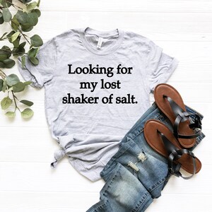 May include: A gray t-shirt with the text "Looking for my lost shaker of salt." printed in black. The shirt is laid out on a white surface with a pair of blue jeans and brown sandals.