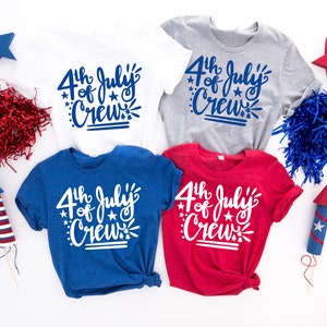 May include: Four t-shirts in white, gray, blue, and red, each with the text "4th of July Crew" in a script font. The shirts are surrounded by patriotic decorations, including stars, paper filler, and toy rockets.