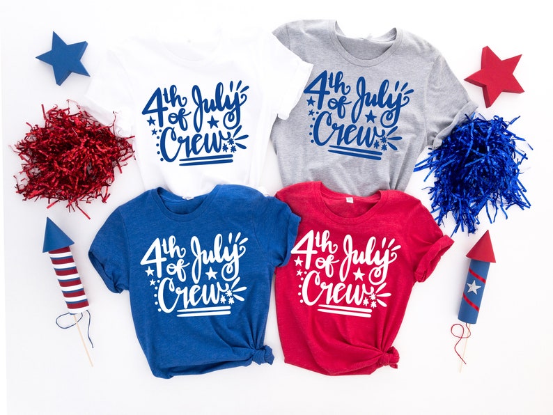 May include: Four t-shirts in white, gray, blue, and red, each with the text "4th of July Crew" in a script font. The shirts are surrounded by patriotic decorations, including stars, paper filler, and toy rockets.