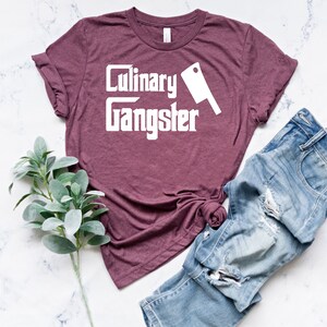 May include: A maroon t-shirt with the text "Culinary Gangster" in white lettering and a white silhouette of a butcher knife.