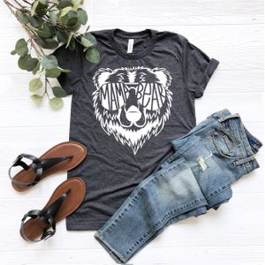 May include: A dark gray t-shirt with a white bear graphic. The bear is wearing sunglasses and the text "Mama Bear" is inside the sunglasses. The t-shirt is laid out on a white wooden surface with a pair of brown sandals and blue jeans.