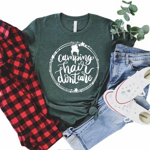 May include: A green t-shirt with the text "Camping hair don't care" in a white circle with stars around it. The shirt is laid out on a white background with a red and black plaid shirt, blue jeans, and white sneakers.