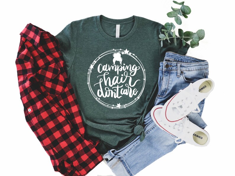 May include: A green t-shirt with the text "Camping hair don't care" in a white circle with stars around it. The shirt is laid out on a white background with a red and black plaid shirt, blue jeans, and white sneakers.