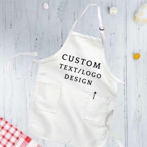May include: White cotton apron with a large pocket and adjustable neck straps. The apron has black text that reads "CUSTOM TEXT/LOGO DESIGN".