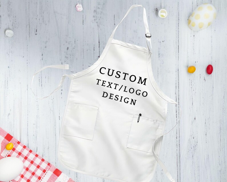 May include: White cotton apron with a large pocket and adjustable neck straps. The apron has black text that reads "CUSTOM TEXT/LOGO DESIGN".