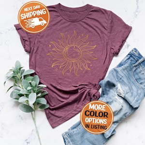 May include: A maroon t-shirt with a gold sun and moon design. The design features a stylized sun with rays and a crescent moon inside. The text "More Color Options in Listing" is on a brown circle.