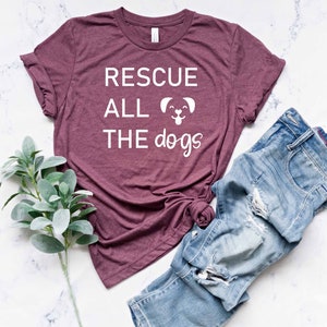 K&ouml;nnte beinhalten: Ein maroonfarbenes T-Shirt mit dem Text "RESCUE ALL THE dogs" in Wei&szlig; mit einer kleinen Hundgrafik. Das Shirt liegt flach auf einer wei&szlig;en Marmorfl&auml;che mit einer blauen Jeans und einem Zweig Gr&uuml;n.