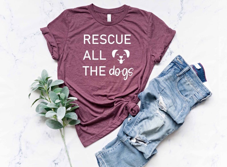 K&ouml;nnte beinhalten: Ein maroonfarbenes T-Shirt mit dem Text "RESCUE ALL THE dogs" in Wei&szlig; mit einer kleinen Hundgrafik. Das Shirt liegt flach auf einer wei&szlig;en Marmorfl&auml;che mit einer blauen Jeans und einem Zweig Gr&uuml;n.