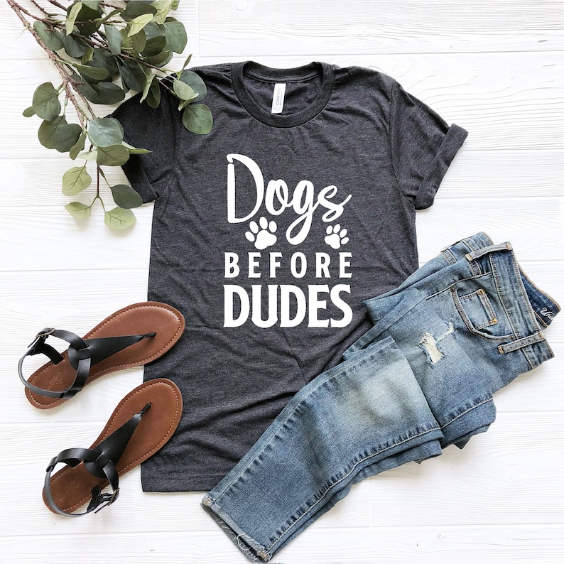 May include: A dark grey t-shirt with white text that reads "Dogs Before Dudes" with two paw prints above the word "Before". The shirt is laid flat on a white wooden surface with a pair of brown sandals and blue jeans.