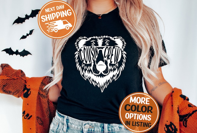 May include: Black t-shirt featuring a white bear graphic wearing sunglasses. The sunglasses spell out "COUSIN CREW".