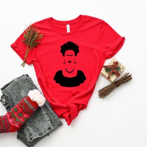 以下が含まれることがあります： 前面にフリーダ・カーロの黒いシルエットがプリントされた赤いTシャツ
