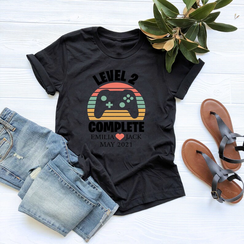 May include: Black t-shirt with a retro sunset graphic and a video game controller. The text "Level 2 Complete" is printed on the shirt, along with the names "Emilia" and "Jack" and the date "May 2021".
