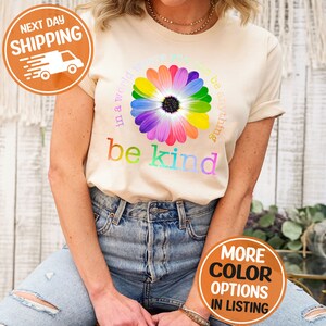Puede incluir: Camiseta color crema con un dise&ntilde;o de margarita arco&iacute;ris y las palabras "in a world where you can be anything, be kind". La camiseta tiene cuello redondo y mangas cortas. La imagen tambi&eacute;n incluye un gr&aacute;fico de env&iacute;o y opciones de color.