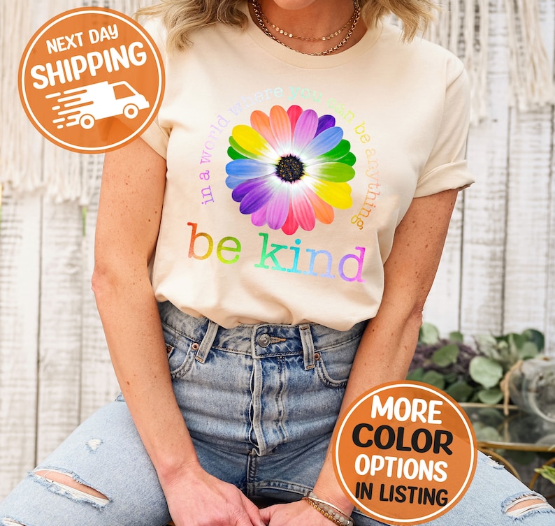 Puede incluir: Camiseta color crema con un dise&ntilde;o de margarita arco&iacute;ris y las palabras "in a world where you can be anything, be kind". La camiseta tiene cuello redondo y mangas cortas. La imagen tambi&eacute;n incluye un gr&aacute;fico de env&iacute;o y opciones de color.