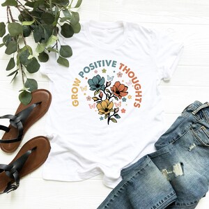 May include: White t-shirt with a floral design and the text "Grow Positive Thoughts" in a circular arc around the flowers.