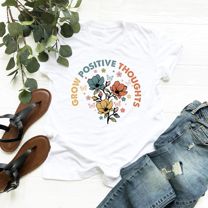 May include: White t-shirt with a floral design and the text "Grow Positive Thoughts" in a circular arc around the flowers.