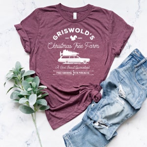 May include: A maroon t-shirt with a white graphic design that says "Griswold's Christmas Tree Farm" with a squirrel, a station wagon with a Christmas tree on top, and the text "A Real Beaut Guaranteed" and "Free Squirrel With Purchase".