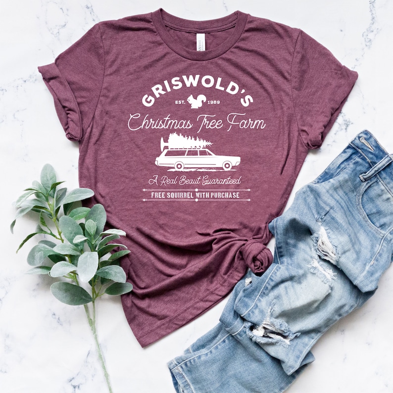May include: A maroon t-shirt with a white graphic design that says "Griswold's Christmas Tree Farm" with a squirrel, a station wagon with a Christmas tree on top, and the text "A Real Beaut Guaranteed" and "Free Squirrel With Purchase".