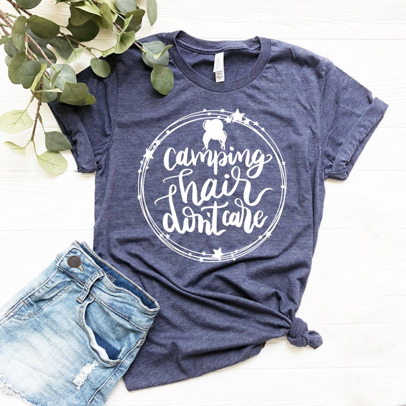 May include: A navy blue t-shirt with a white circle design featuring the text "camping hair don't care" and a silhouette of a woman with messy hair. The shirt is laid flat on a white wooden surface.