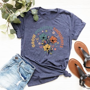 May include: A navy blue t-shirt with a floral design and the text "Grow Positive Thoughts" in a circle around the flowers.