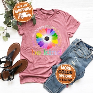 Puede incluir: Una camiseta rosa polvorienta con un gr&aacute;fico de margarita arco&iacute;ris y las palabras "be kind". La camiseta est&aacute; combinada con vaqueros azules y sandalias negras. El texto "el mundo donde puedes ser cualquier cosa" tambi&eacute;n est&aacute; en la camiseta.
