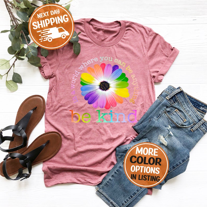 Puede incluir: Una camiseta rosa polvorienta con un gr&aacute;fico de margarita arco&iacute;ris y las palabras "be kind". La camiseta est&aacute; combinada con vaqueros azules y sandalias negras. El texto "el mundo donde puedes ser cualquier cosa" tambi&eacute;n est&aacute; en la camiseta.