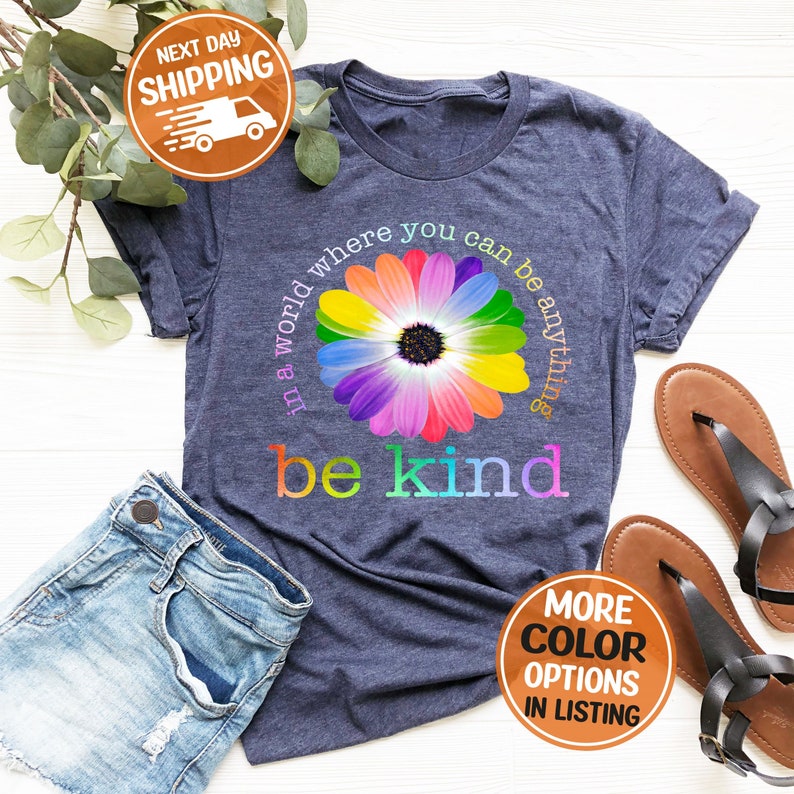 Puede incluir: Camiseta azul marino con un dise&ntilde;o de margarita colorida y las palabras "in a world where you can be anything, be kind". La camiseta se combina con pantalones cortos de mezclilla y sandalias. La camiseta tiene cuello redondo y mangas cortas.