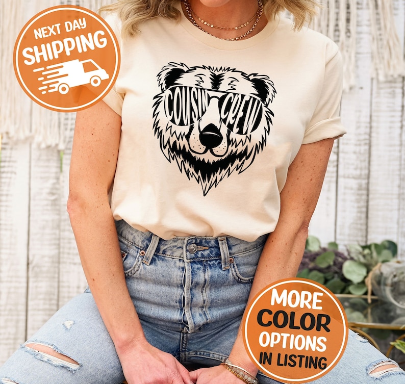 May include: A beige t-shirt featuring a black bear wearing sunglasses. The sunglasses have the words "COUSIN CREW" written on them.