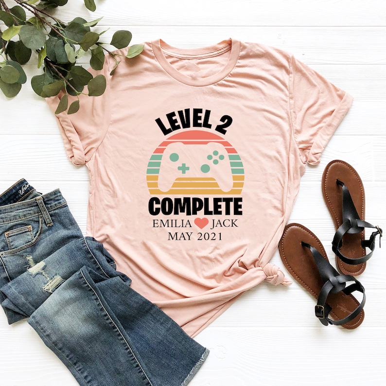 May include: A pink t-shirt with a retro-style video game controller graphic and the text "Level 2 Complete" with the names "Emilia" and "Jack" and the date "May 2021".