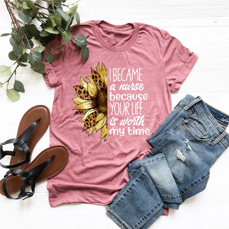 Op de afbeelding: Een roze T-shirt met een zonnebloemdessin en de tekst "Ik ben verpleegster geworden omdat je leven mijn tijd waard is".