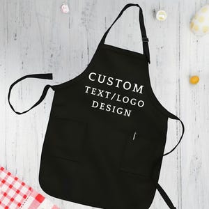 May include: Black apron with adjustable straps and a large pocket. The apron has white text that reads "CUSTOM TEXT/LOGO DESIGN".
