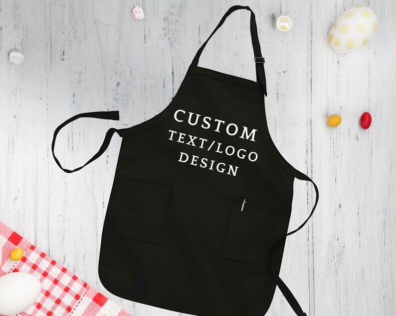 May include: Black apron with adjustable straps and a large pocket. The apron has white text that reads "CUSTOM TEXT/LOGO DESIGN".