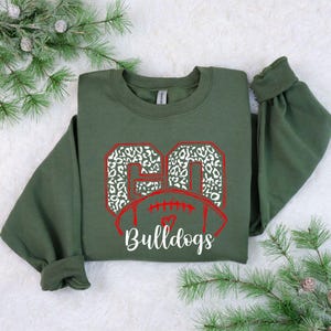 May include: Green sweatshirt with a red and white football design. The football has the number 60 in white with a leopard print pattern. The text "Bulldogs" is below the football.