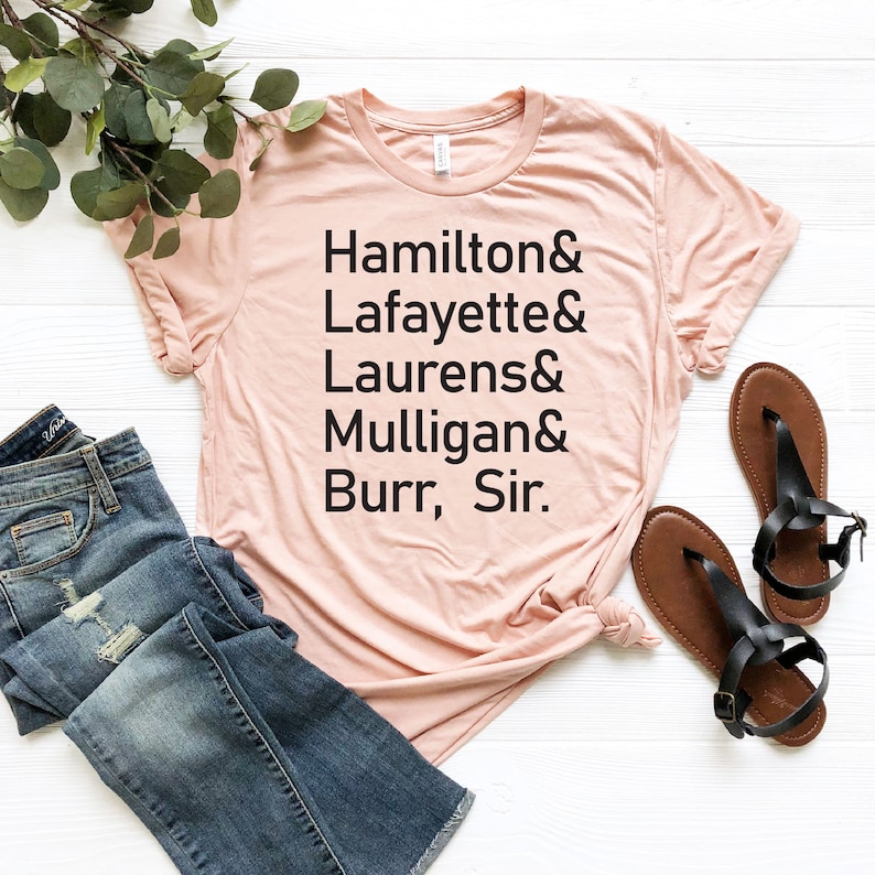 Op de afbeelding: Een lichtroze T-shirt met zwarte tekst die "Hamilton & Lafayette & Laurens & Mulligan & Burr, Sir." luidt.