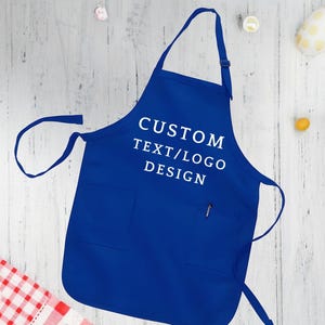 May include: A royal blue apron with a large pocket and adjustable straps. The apron has white text that reads "CUSTOM TEXT/LOGO DESIGN".