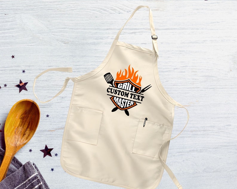 Puede incluir: Un delantal de algod&oacute;n beige con un bolsillo grande y dos bolsillos m&aacute;s peque&ntilde;os. El delantal tiene un gr&aacute;fico negro y naranja que dice "Grill Custom Text Master" con un dise&ntilde;o de llama.